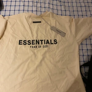 Beige Essentials Fear of God t-shirt - Snygg beige t-shirt från Essentials Fear of God med svart logga tryckt på bröstet. Klassisk passform med rund hals och korta ärmar. Tillverkad i mjuk bomull, perfekt för en clean och stilren look.