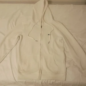 Vit hoodie från Lacoste med dragkedja storlek s - Säljer en stilren vit hoodie från Lacoste med klassisk krokodillogga på bröstet. Hoodien har dragkedja, snörning i huvan och två stora fickor framtill. Perfekt för dig som gillar en clean och sportig look.