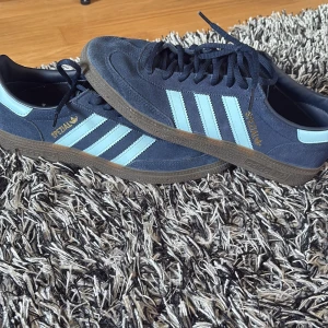Adidas Spezial sneakers marinblå mocka - Säljer ett par Adidas Spezial sneakers i marinblå mocka med ljusblå ränder och detaljer. Skorna har klassisk brun gummisula, snörning och rund tå. Ikonisk retrostil med guldfärgad Spezial-logga på sidan. Perfekta för dig som gillar streetwear och snygga detaljer.