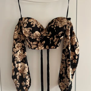 Blommig svart korsettopp med puffärm - Supersnygg svart korsettopp med beige blommönster och ballongärmar. Toppen har smala axelband, markerad byst och långa band framtill för knyt. Perfekt för dig som vill sticka ut med en trendig och feminin look.