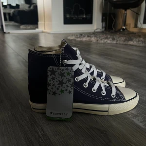 Converse Chuck Taylor All Star High - Klassiska Converse Chuck Taylor All Star high-top sneakers i mörkblå canvas med vita snören och kontrasterande vita sömmar. Skorna har rund tå, platt sula och den ikoniska All Star-loggan på sidan. Perfekta för en avslappnad och cool streetstil.
