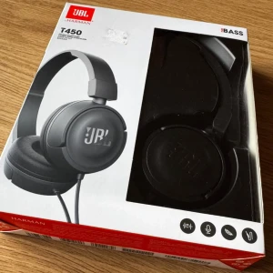 JBL T450 - JBL T450 är lätta, hopfällbara over-ear hörlurar med JBL Pure Bass-ljud, inbyggd mikrofon och fjärrkontroll. Kabeln är platt och trasselfri. Produkten är i originalförpackning och ser oanvänd ut, perfekt för musik och samtal på språng.