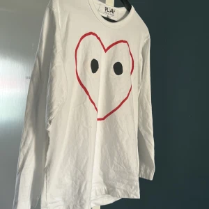 Vit långärmad tröja Comme des Garçons - Säljer en vit långärmad tröja från Comme des Garçons PLAY med det ikoniska röda hjärtat med ögon tryckt på bröstet. Tröjan är i mjuk bomull och har en avslappnad passform, perfekt för en cool och stilren look.