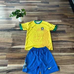 Brasilien Pelé 10 Nike fotbollströja - Säljer en klassisk Brasilien fotbollströja från Nike med Pelé och nummer 10 på ryggen. Tröjan är gul med gröna detaljer och blå Nike-logga, tillverkad i Dri-Fit material. Matchande blå shorts med gul Nike-logga och Brasilien-emblem ingår.