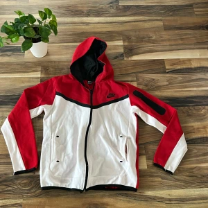 Nike röd och vit vindjacka med huva - Nike vindjacka i rött och vitt med svart detalj och dragkedja. Jackan har huva, svarta detaljer, fickor med dragkedja och en extra ficka på ärmen. Perfekt för dig som gillar sportig stil och vill sticka ut med färg.