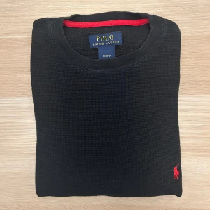 Ralph Lauren Stickad Tröja Svart S - Svart långärmad tröja från Polo Ralph Lauren i 100% bomull. Tröjan har en diskret struktur, rund halsringning med röd detalj och den klassiska röda broderade loggan på bröstet. Perfekt för dig som gillar stilrena och tidlösa plagg.
