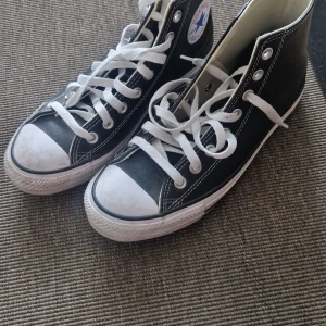 Svarta Converse All Star high-top - Klassiska svarta Converse All Star high-top sneakers i skinn med vita snören och gummisula. Ikonisk logga på sidan och kontrasterande vita sömmar. Perfekta för dig som vill ha en tidlös och cool stil.