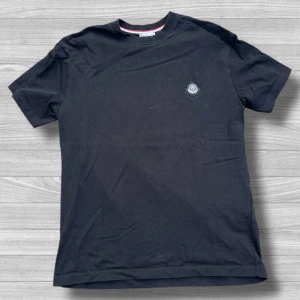 Svart t-shirt från Moncler, storlek S - Svart t-shirt från Moncler i mjuk bomull, med klassisk rund hals och korta ärmar. Diskret Moncler-logga på bröstet och snygg detalj med röd och vit rand på insidan av kragen. Perfekt för dig som gillar stilrena plagg med exklusiv känsla.