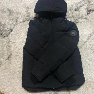 Svart macmilan från Canada Goose - Säljer en svart dunjacka från Canada Goose med huva och klassisk patch på ärmen. Jackan har dragkedja framtill, flera fickor och är riktigt varm och skön för kalla dagar. Perfekt för dig som vill ha en stilren och funktionell vinterjacka.. Accepterar inget under 2650kr