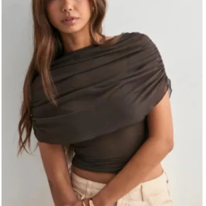 Svart croppad mesh topp - Säljer en svart croppad topp i mesh med omlottkänsla och draperad effekt över axlarna. Toppen är tight och har en unik offshoulder-design som ger en cool och trendig look. Perfekt för dig som vill sticka ut lite extra.