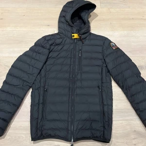 Svart dunjacka från Parajumpers - Svart dunjacka från Parajumpers med huva och klassisk patch på ärmen. Jackan har quiltad design, flera dragkedjeförsedda fickor och en gul detalj vid halsen. Perfekt för kalla dagar och riktigt stilig!