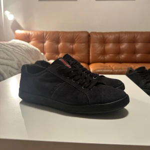 Svarta mockasneakers från Prada - Snygga svarta sneakers från Prada i mocka med diskret logga på sidan och röd detalj med Prada-tryck på plösen. Skorna har rund tå, platt sula och klassisk snörning. Perfekta för dig som gillar stilrena och exklusiva sneakers.