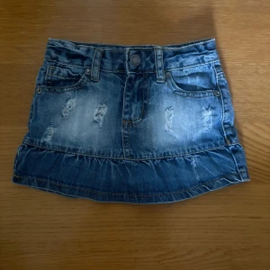 Blå jeanskjol med volangkant - Snygg blå jeanskjol med slitna detaljer och volangkant längst ner. Kjolen har klassiska fickor fram och bak, bälteshällor och knappstängning. Perfekt för dig som gillar en cool och avslappnad stil.