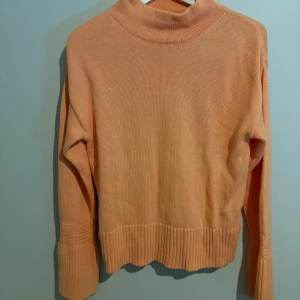Persikofärgad stickad tröja från Pelle P - Säljer en persikofärgad stickad tröja från Pelle P med hög krage och breda ribbade muddar vid ärmslut och nederkant. Tröjan har lång ärm och är i mjukt material, perfekt för kyliga dagar. Enkel och stilren design som är lätt att matcha.