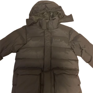 Svart pufferjacka från Helly Hansen - Säljer en svart pufferjacka från Helly Hansen med huva och stora fickor framtill. Jackan har Helly Hansen-logga på bröstet och nedtill, dragkedja och är vadderad för extra värme. Perfekt för kalla dagar och riktigt snygg streetstil. Till pojkar storlek 164 