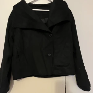 Svart kappa från Gina Tricot - Stilren svart kappa från Gina Tricot med bred krage och två knappar framtill. Jackan har en rak, något croppad passform och är helfärgad utan mönster. Perfekt för dig som gillar minimalistisk och klassisk stil.