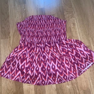 Mönstrad bandeau tubtopp i rosa och lila - Säljer en bandeau tubtopp med elastisk smock upptill och ett färgstarkt mönster i rosa, lila och rött. Toppen är ärmlös och har en luftig, utsvängd passform nedtill. Perfekt för varma dagar när du vill sticka ut med en cool look.