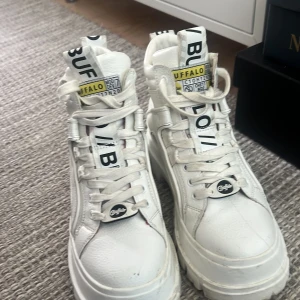 Buffalo vita chunky sneakers - Buffalo vita chunky sneakers med hög sula och grovt mönstrad yttersula. Skorna har snörning, detaljerade logotyper på plösen och sidan samt insida med gul och rutig detalj. Tillverkade i syntetmaterial med läderlook och cool streetstil.