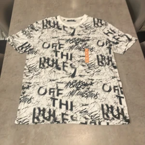 Vit t-shirt med graffiti från XSIDE - Säljer en vit t-shirt från XSIDE i storlek M med svart graffiti-inspirerat tryck och text som 'OFF THE RULES' och 'NO QUESTIONS'. T-shirten har korta ärmar och en relaxed passform. Perfekt för dig som gillar streetstyle och unika prints.