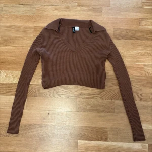 Brun ribbad croppad tröja  - Säljer en brun ribbad croppad tröja från Divided med v-ringning och krage. Tröjan har långa ärmar och är i ett mjukt stickat material som sitter snyggt och bekvämt. Perfekt för lager-på-lager och snygg till jeans eller kjol.