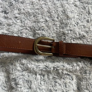 Brunt skärp med guldfärgat spänne - Snyggt brunt bälte i skinn med klassiskt guldfärgat spänne. Bältet har en stilren design med lätt struktur i materialet och raka kanter. Perfekt accessoar för att lyfta din outfit med en tidlös touch.
