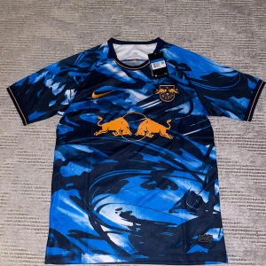 RB Leipzig blå Nike fotbollströja - Snygg RB Leipzig fotbollströja från Nike med blått och svart camouflage-mönster och orange detaljer. Tröjan har klubbmärke och Red Bull-logga på bröstet samt rund halsringning. Tillverkad i lätt och ventilerande material med Dri-FIT-teknologi.