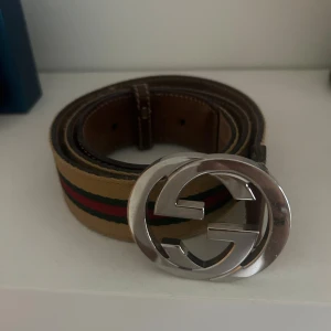 Bälte med GG-spänne - Snyggt bälte med klassiskt GG-spänne i silverfärgad metall. Bältet har en gul bas med gröna och röda ränder längs mitten och insida i brunt skinn. Perfekt accessoar för att lyfta din outfit med en lyxig touch.