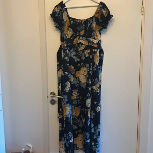 Blommig långklänning med puffärm - Supersöt långklänning med marinblå botten och stora gula och beige blommor. Klänningen har puffiga korta ärmar och markerad midja. Perfekt för sommarens alla tillfällen och ger en romantisk vibe.
