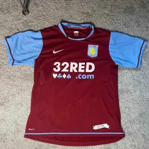 Aston Villa fotbollströja 2007/08 - Aston Villa fotbollströja från Nike i vinrött med ljusblå ärmar och detaljer. Klassisk klubbsköld på bröstet, sponsortryck och nummer 13 med initialer TJ på ryggen. Tillverkad i lätt och ventilerande polyester med NikeFIT-teknologi.