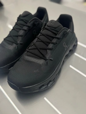 Svarta sneakers från On Running - Snygga helsvarta sneakers från On Running med modern design och ventilerande meshmaterial. Skorna har rund tå, snörning och en unik sula med hål för extra komfort och stil. Perfekta för dig som gillar sportig och clean look.