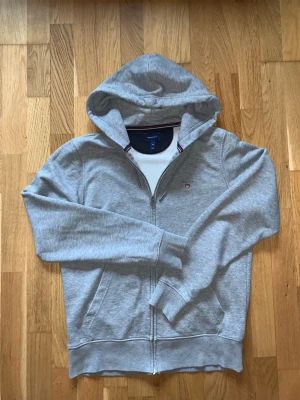 Grå zip hoodie från Gant - Snygg grå hoodie från Gant med dragkedja framtill och två fickor. Klassisk huva och diskret Gant-logga på bröstet. Muddar vid ärmslut och nederkant. Tillverkad i mjukt bomullsmaterial för skön känsla. Perfekt för en avslappnad och stilren look. Vid frågor är det bara att höra av sig!