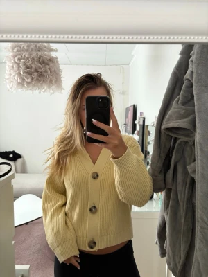 gul ribbstickad kofta med stora knappar - Trendig gul kofta med ribbstickat mönster och tre stora bruna knappar framtill. Koftan har v-ringning, långa ärmar och breda muddar. Perfekt att slänga på sig över en t-shirt eller linne för en avslappnad look.