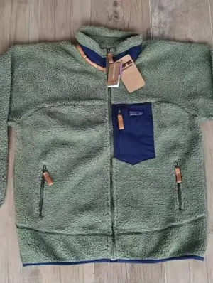 Grön teddyjacka från Patagonia - Mysig grön teddyjacka från Patagonia med hel dragkedja framtill och kontrasterande mörkblå bröstficka. Jackan har två sidofickor med dragkedja och detaljer i läder på dragkedjorna. Hög krage och avslut i blått nertill ger en clean look.