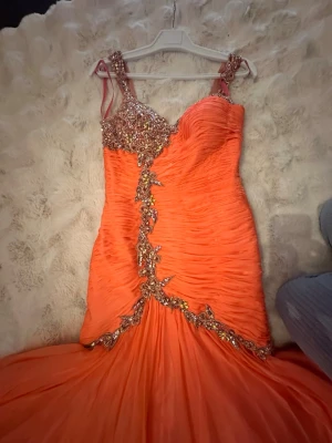 Lång korallfärgad balklänning  - Lång, korallfärgad balklänning med vackra detaljer. Klänningen har en figurnära passform upptill och en utsvängd kjol nedtill. Den är dekorerad med strass och paljetter runt bysten och längs framsidan. Skicket enligt bilderna  Koral färg . Använd endast en gång och är ej kemtvättad