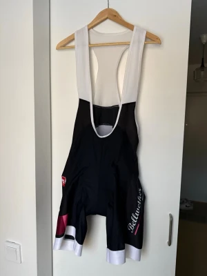 Bellwether cykelbyxa med hängslen - Snygga cykelbyxor med hängslen från Bellwether. Svart bas med vita axelband och detaljer i vitt, rosa och blått. Tight passform och korta ben, perfekt för cykling. Materialet är stretchigt och andas bra.