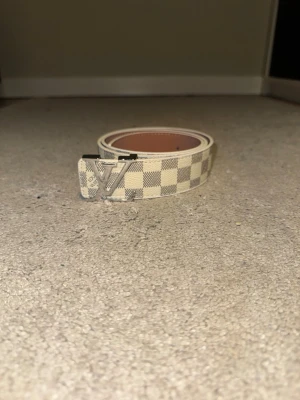 Rutmönstrat bälte med metallspänne - Oanvänt bälte i ljust rutmönster med kontrasterande metallspänne i svart/silverfärg. Klassisk och tidlös design, i nyskick.