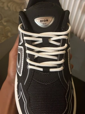 Svarta Sneakers - Säljer ett par svarta  sneakers med vit sula och reflekterande detaljer på sidorna. Skorna har vit snörning, meshpaneler. Snygg och modern design som sticker ut.