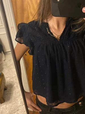 Mörkblå blus med volangärmar - Söt mörkblå blus med korta volangärmar och v-ringning. Blusen har ett diskret prickigt mönster och är lätt transparent med luftig passform. Perfekt till jeans eller kjol för en trendig look.