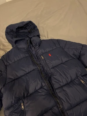 Mörkblå pufferjacka från Polo Ralph Lauren - Snygg mörkblå pufferjacka från Polo Ralph Lauren med huva och dragkedja framtill. Jackan har det klassiska röda Polo-logotypen broderad på bröstet och flera praktiska fickor med dragkedja. Perfekt för kalla dagar och riktigt stilren.