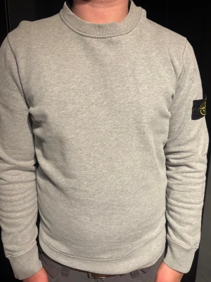 Grå sweatshirt från Stone Island - Säljer en stilren grå sweatshirt från Stone Island med klassisk rund hals och ribbade muddar. Tröjan har den ikoniska svarta patchen med gul kompasslogga på vänster ärm. Tillverkad i mjuk bomullsblandning för skön känsla och enkel passform. Tröjan är äkta och i super fint skick.