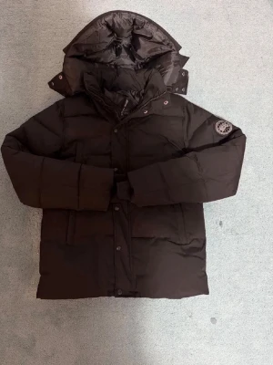 Svart dunjacka från Canada Goose - Snygg svart dunjacka från Canada Goose med huva och klassisk patch på ärmen. Jackan har lång ärm, dragkedja och knappar framtill samt en quiltad design som håller dig varm under kalla dagar. Perfekt för dig som vill ha både stil och funktion. Säljer den då den ena dragkedjan har gått av och gått i sönder men man kan ju altid köpa en ny bara och trä på tar ju bara nån sekund pris grå o diskuteras vid snabb affär mvh robin