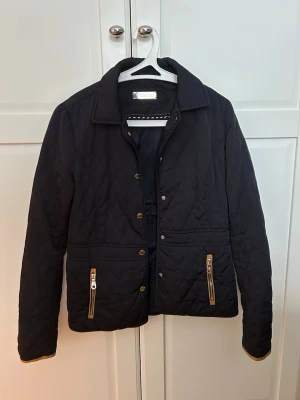 Svart/mörkblå quiltad jacka Massimo Dutti - Stilren svart/ mörkblå quiltad jacka från Massimo Dutti med guldiga dragkedjor på fickorna och knappar framtill. Jackan har klassisk krage och snyggt mönstrad insida. Perfekt för dig som gillar en tidlös look. Den är obetydligt använd, säljs då den är för liten i storlek. 