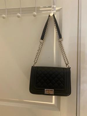 Svart handväska med kedja - Stilren svart handväska med quiltat mönster och silverfärgad kedjerem. Väskan har en rektangulär form, elegant lås framtill och detaljer i metall. Perfekt accessoar för att lyfta din outfit. Är använd men i bra skick! Pris kan alltid diskuteras 🙌🏼