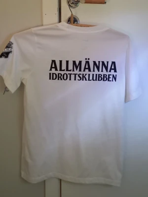 Vit Nike AIK t-shirt med tryck - Vit t-shirt från Nike med svart Swoosh-logga på bröstet och stort 'ALLMÄNNA IDROTTSKLUBBEN'-tryck på ryggen. AIK-märke på ärmen. Klassisk passform och rund hals. Perfekt för dig som gillar sportig stil och AIK. Storlek 158-170