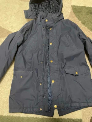 Mörkblå quiltad parkas med huva”pris kan diskuteras” - Snygg mörkblå parkajacka från Stadium med quiltat innerfoder och avtagbar huva. Jackan har dragkedja och knappar framtill samt praktiska fickor med lock och tryckknappar. Perfekt för kyliga dagar och enkel att matcha med olika outfits.