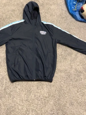 Svart vindjacka från North Sails - Snygg svart vindjacka från Norra läktaren Malmö ff med huva och dragkedja framtill. Jackan har vita och ljusblå sopar längs ärmarna och två sidofickor. Perfekt för blåsiga dagar och enkel att matcha till olika outfits.