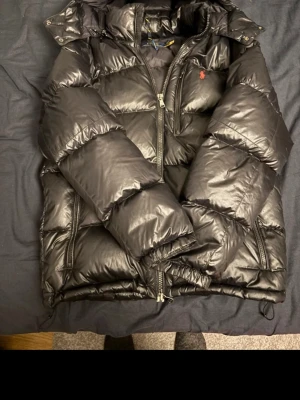 Svart dunjacka från Polo Ralph Lauren - Säljer en svart dunjacka från Polo Ralph Lauren med klassisk pufferlook och röd logga på bröstet. Jackan har huva, dragkedja framtill och två sidofickor med dragkedja. Perfekt för kalla dagar och riktigt snygg till alla outfits.