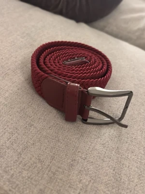 Vinrött flätat bälte med metallspänne - Snyggt vinrött bälte i flätad textil med detaljer i konstläder och ett stilrent metallspänne. Perfekt accessoar för att lyfta din outfit med en färgklick. Passar både till jeans och chinos för en trendig look.