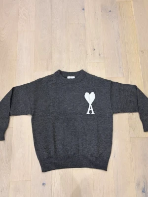 Mörkgrå stickad tröja från Ami Paris - Mörkgrå stickad tröja från Ami Paris med rund halsringning och ribbade muddar. På bröstet finns ett vitt hjärta med bokstaven A. Perfekt för dig som gillar stilrena och trendiga plagg.