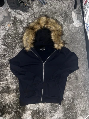 Svart hoodie med fuskpäls på huvan - Svart hoodie med dragkedja från H&M. Huvan är fodrad med mjuk fuskpäls i bruna toner som ger en snygg kontrast. Klassisk passform med muddar vid ärmslut och nederkant samt två fickor framtill. Perfekt för en chill och trendig look.